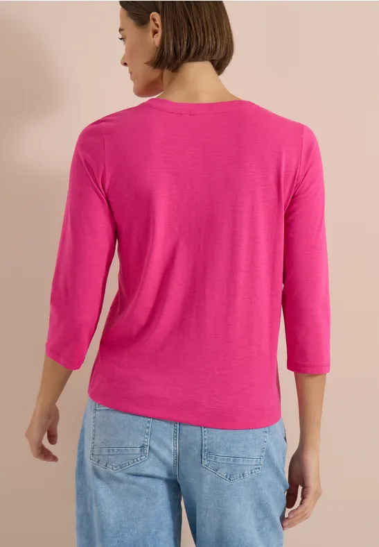 Frau von hinten in pinkem 3/4-Arm-Shirt und blauen Jeans vor beigem Hintergrund.