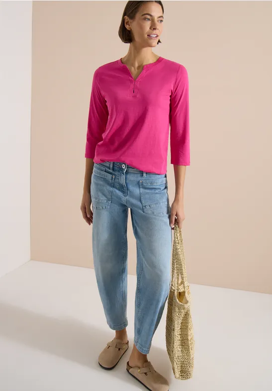 Frau trägt pinkes Henley-Shirt, helle Boyfriend-Jeans, Basttasche und beige Loafers vor hellem Hintergrund.