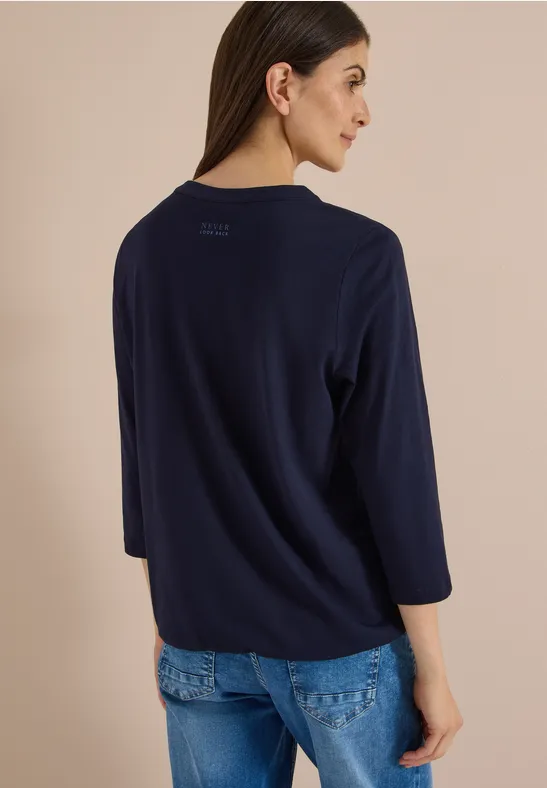 Frau in dunkelblauem Pullover mit 3/4-Ärmeln und Jeans, Rückenansicht vor hellem Hintergrund.