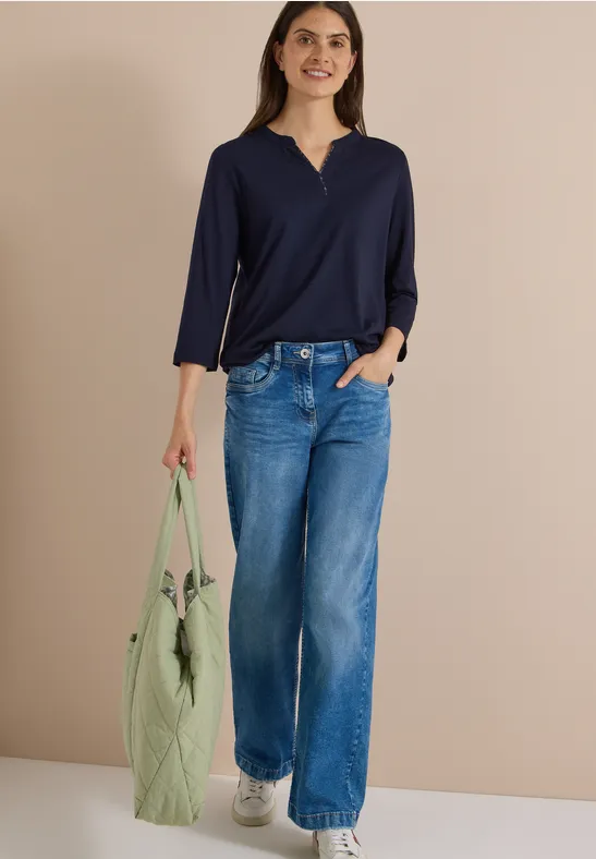 Frau in dunkelblauer Bluse, weiten Jeans und grüner Tasche mit Bommel vor neutralem Hintergrund.