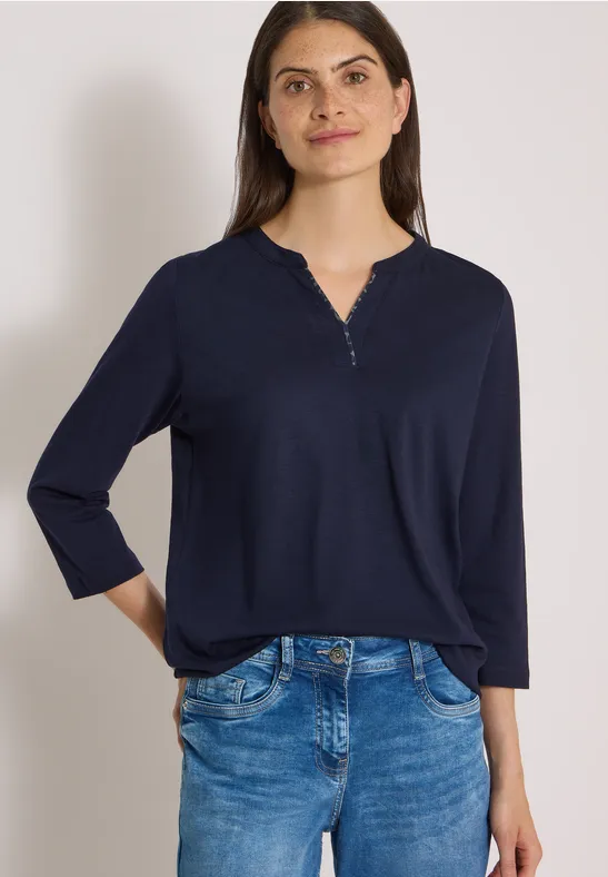 Frau trägt dunkelblaues Shirt mit 3/4-Ärmeln und Jeans vor weißem Hintergrund.