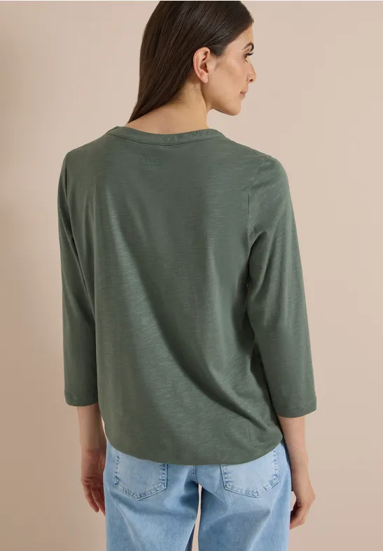 Vrouw van achter gefotografeerd in kaki 3/4-mouwen shirt en lichtblauwe spijkerbroek voor beige achtergrond.