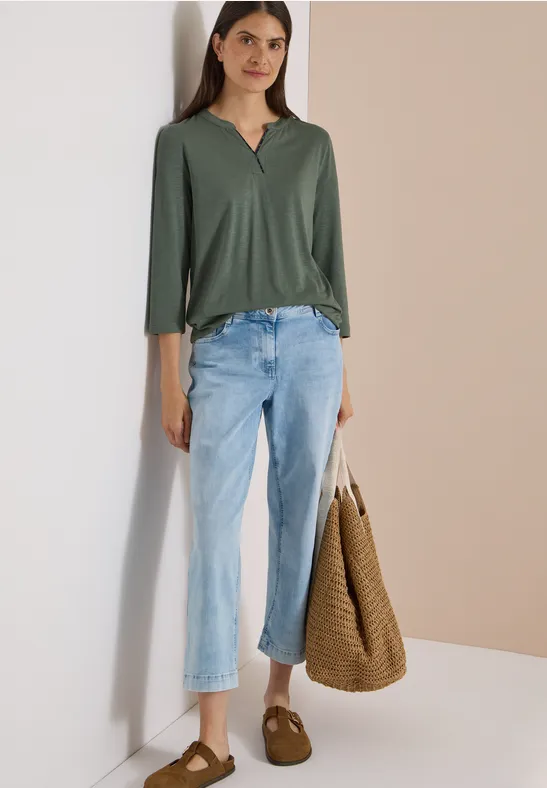 Vrouw in groen shirt en lichte jeans, met bruine slippers en een grote gevlochten tas tegen neutrale achtergrond.
