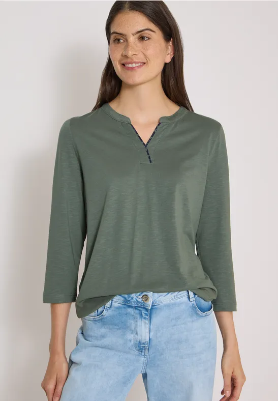 Vrouw glimlachend in groen langarmshirt en blauwe jeans tegen witte achtergrond