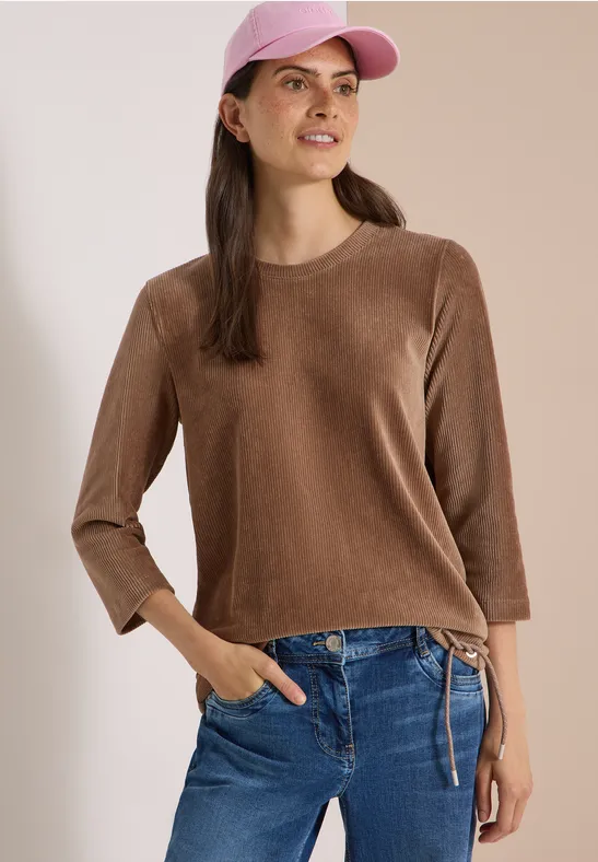 Frau mit braunem 3/4-ärmeligem Pullover, Jeans und rosa Baseballcap lächelt seitlich.
