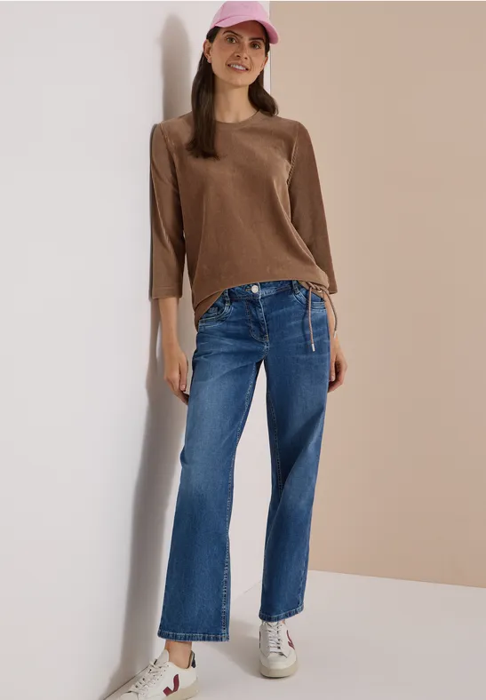 Eine junge Frau lehnt fröhlich an der Wand, trägt braunen Pullover, weite Jeans, weiße Sneaker und rosa Cap.
