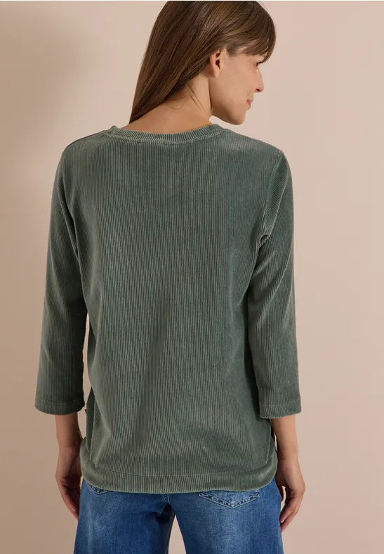 Eine Frau steht mit dem Rücken zur Kamera und trägt ein grünes Langarmshirt mit Jeans.