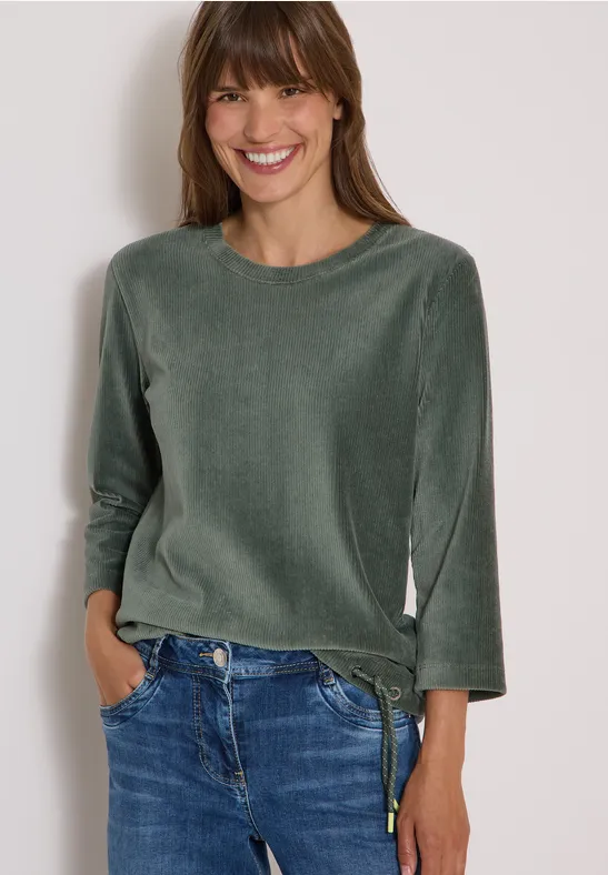 Eine lächelnde Frau in grünem Samtoberteil und blauen Jeans, die Hände in den Taschen.