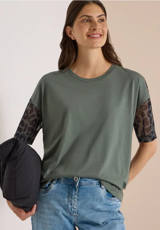Jonge vrouw in groen T-shirt met halfdoorzichtige, motiefmouwen en spijkerbroek, lacht en houdt een jas vast.
