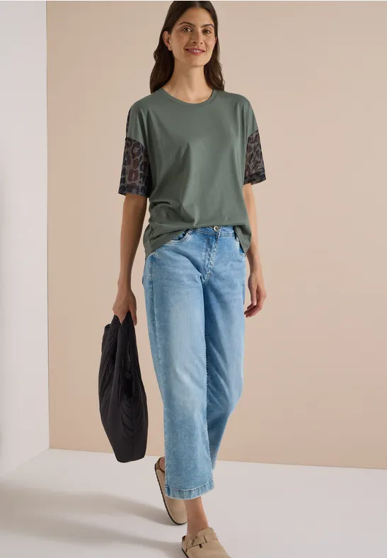 Vrouw draagt olijfgroen shirt met kantmouwen, wijde lichtblauwe jeans en een zwarte tas.