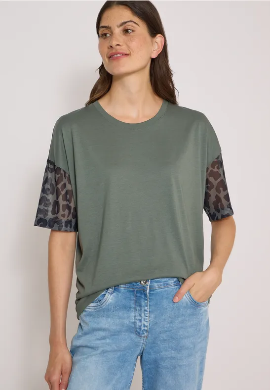 Vrouw in olijfgroen shirt met luipaardprintmouwen en spijkerbroek, hand nonchalant in de zak.