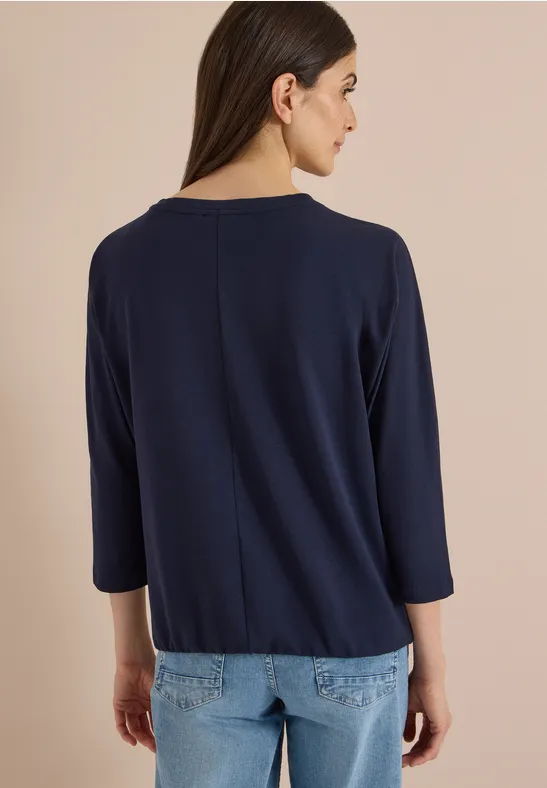 Frau von hinten in marineblauem 3/4-Arm-Oberteil vor neutralem Hintergrund.