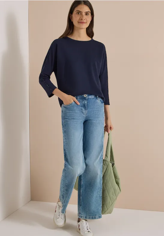 Junge Frau mit dunkelblauem Langarmshirt, hellen Schlagjeans, weißen Sneakern und grüner Tasche vor beigem Hintergrund.