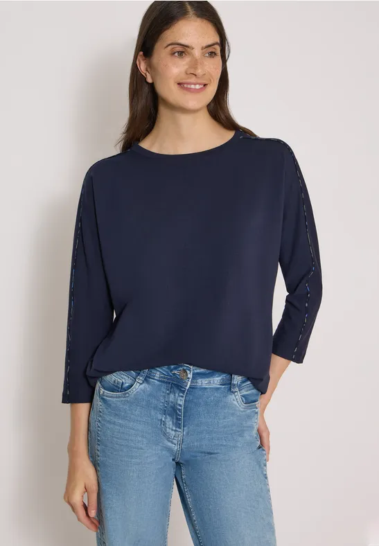 Eine lächelnde Frau in dunkelblauem Dreiviertelarm-Shirt und Jeans vor weißem Hintergrund.