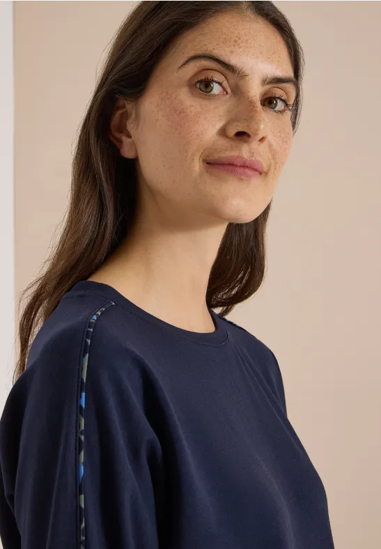 Eine Frau in dunklem Sweatshirt mit dezenten Schulterdetails blickt ruhig zur Kamera.