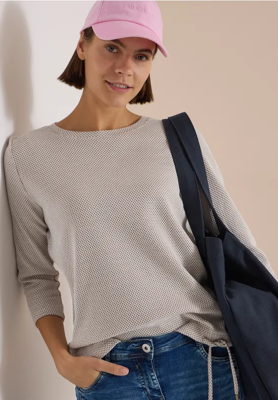 Frau in beiger Longsleeve, Jeans, rosafarbenem Cap und blauer Tasche lehnt an Wand.