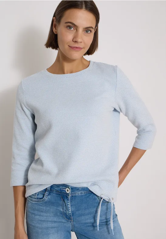 Frau mit schulterlangem Haar in hellblauem Shirt und Jeans vor neutralem Hintergrund.