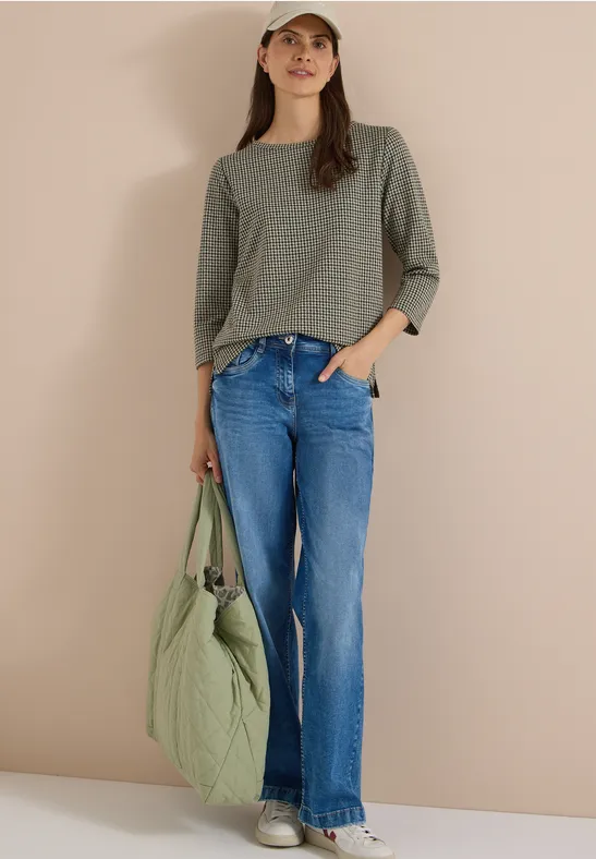 Frau trägt grünes Longsleeve, Schlagjeans und hält eine gesteppte grüne Tasche.