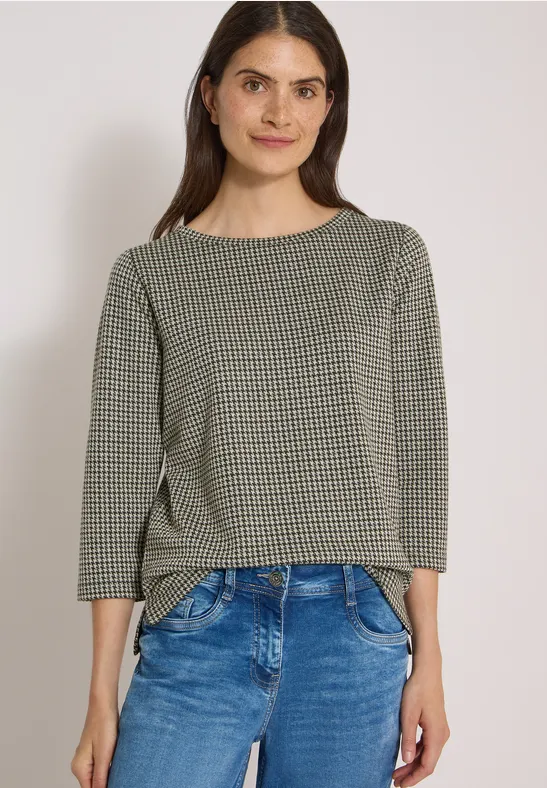 Frau in grauem Musterpullover und blauen Jeans, steht vor neutralem Hintergrund.