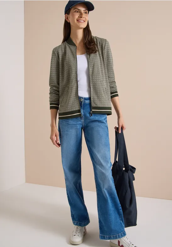 Eine junge Frau in Jeans, weißem Shirt, khakifarbener Jacke, Sneakers und Cap trägt eine große blaue Tasche.