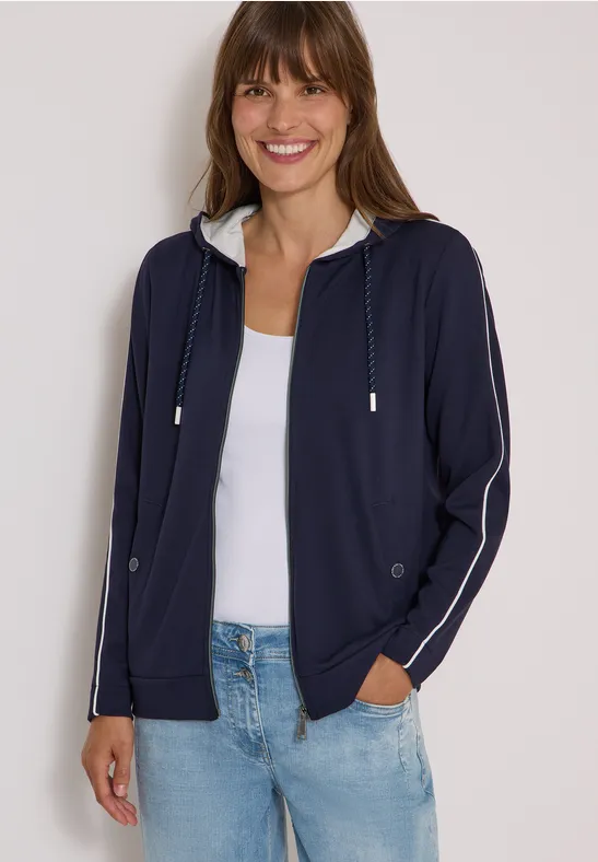 Frau in dunkelblauer Kapuzenjacke, weißem Top und Jeans, lächelnd.