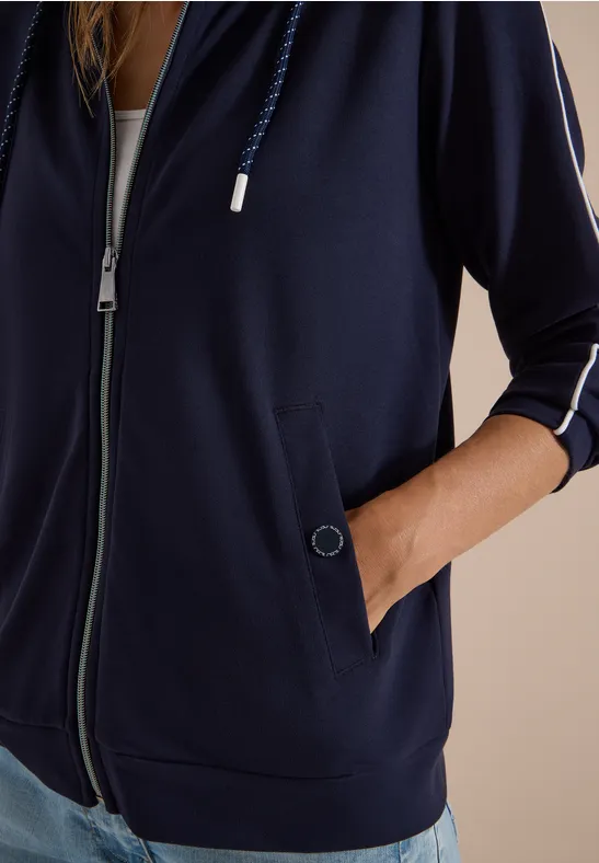Eine Person trägt einen dunkelblauen Zip-Hoodie mit Ärmelnstreifen und steckt die Hand in die Tasche.
