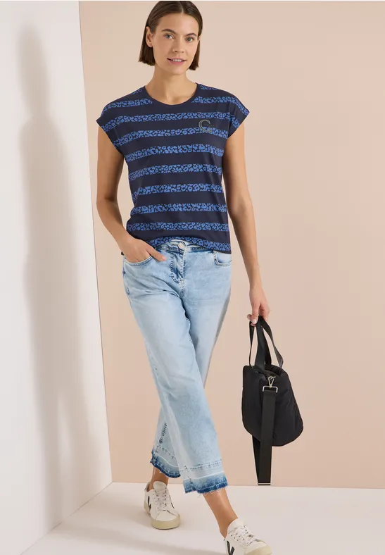 Frau trägt gestreiftes blaues T-Shirt, helle Jeans, weiße Sneaker und hält eine schwarze Tasche.