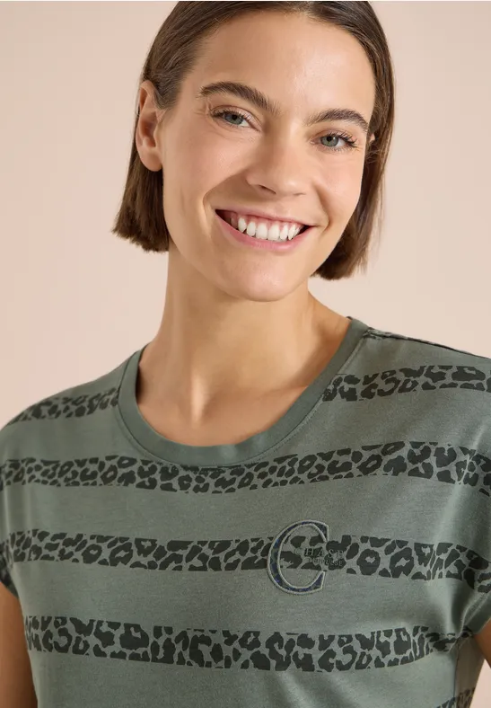 Person mit kurzem Haar trägt grünes T-Shirt mit schwarzen Leopardenstreifen und Kreislogo vor hellem Hintergrund.