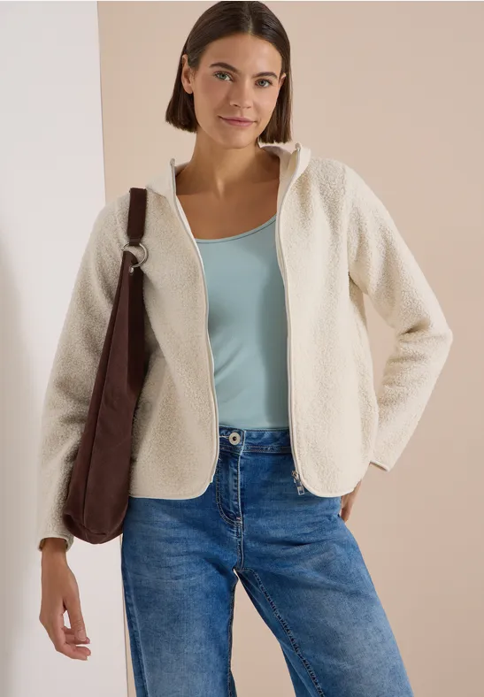 Frau trägt cremefarbene Teddyfleece-Jacke mit Kapuze über türkisfarbenem Top und blauen Jeans, braune Schultertasche.