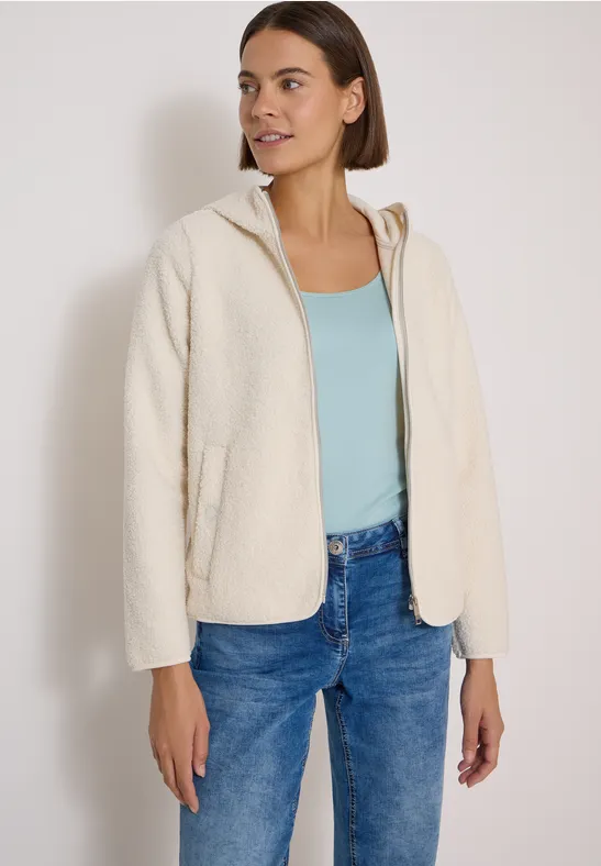 Eine Frau trägt eine cremefarbene Fleecejacke mit Kapuze, ein hellblaues Top und blaue Jeans.
