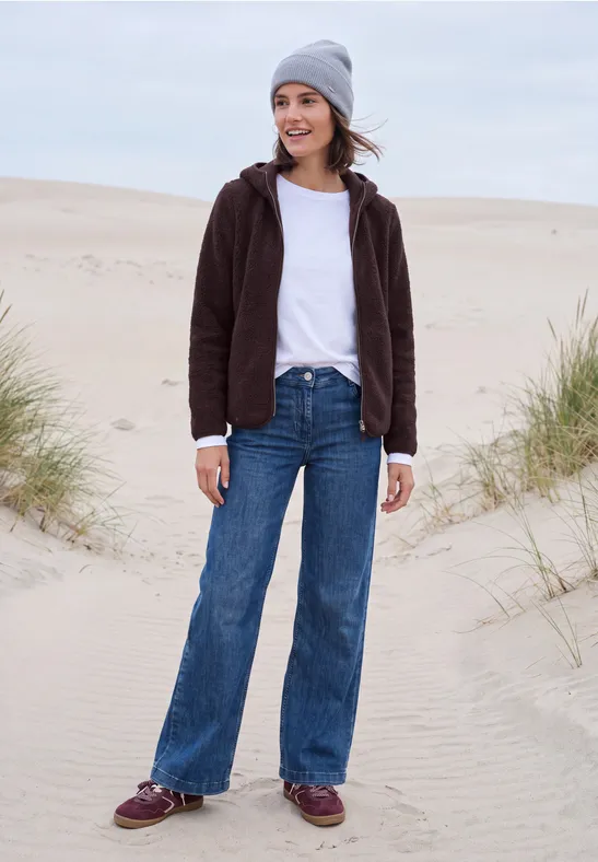 Junge Frau in grauer Mütze, brauner Jacke und weitem Jeans steht lächelnd auf sandigen Dünen.