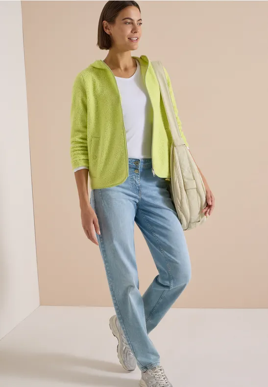 Model in weißem Tanktop, hellgrüner Kapuzenjacke, blauen Jeans, weißen Sneakers und großer Stepp-Tasche.