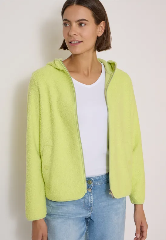 Junge Frau in hellgrüner Fleecejacke mit Kapuze über weißem Top und Jeans.