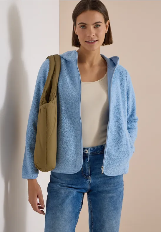 Frau trägt hellblauen Fleece-Pullover mit Kapuze, weißes Top, Jeans und olivgrüne Umhängetasche.