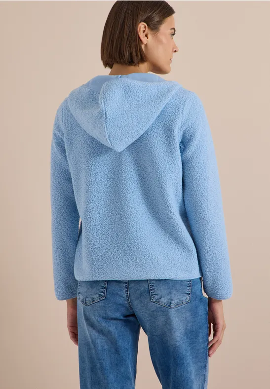 Eine Frau von hinten in einer hellblauen, flauschigen Kapuzenjacke und Jeans vor beigem Hintergrund.