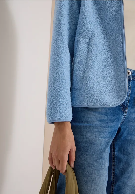 Teilansicht einer Person in Jeans, flauschiger hellblauer Jacke und grüner Tasche.