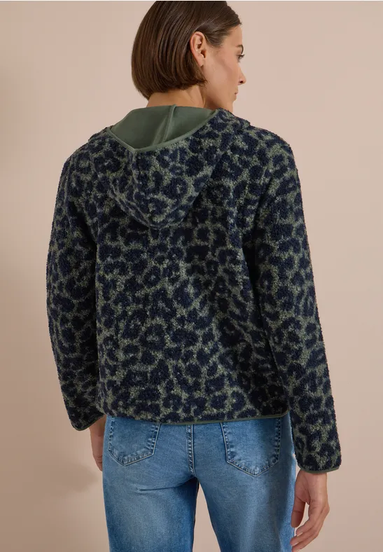 Rückansicht einer Frau in einer blau-grünen gemusterten Fleecejacke mit Kapuze und Jeans vor beigem Hintergrund.