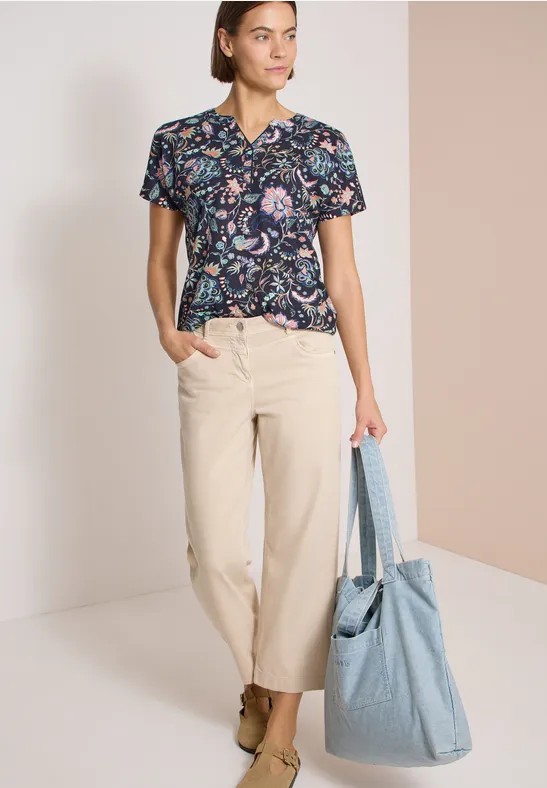 Frau trägt geblümtes T-Shirt, beige Hose und hält eine große hellblaue Tasche.