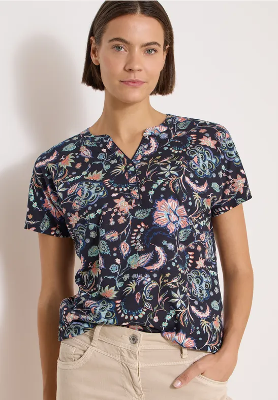 Frau trägt kurzärmeliges dunkelblaues T-Shirt mit buntem Blumen-Paisley-Muster und helle Hose, Hand in Tasche.