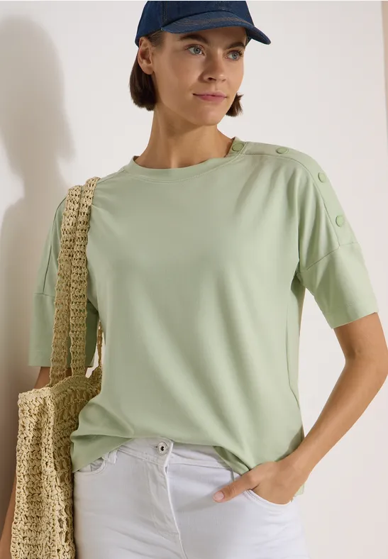 Femme en t-shirt vert menthe, pantalon blanc, sac en paille et casquette devant un mur clair.