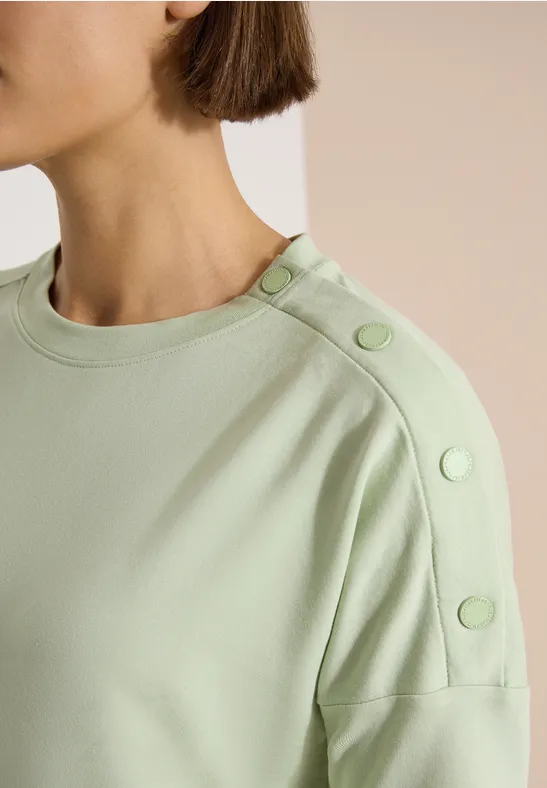 Gros plan sur un haut vert pâle à col rond avec des boutons décoratifs épaule.