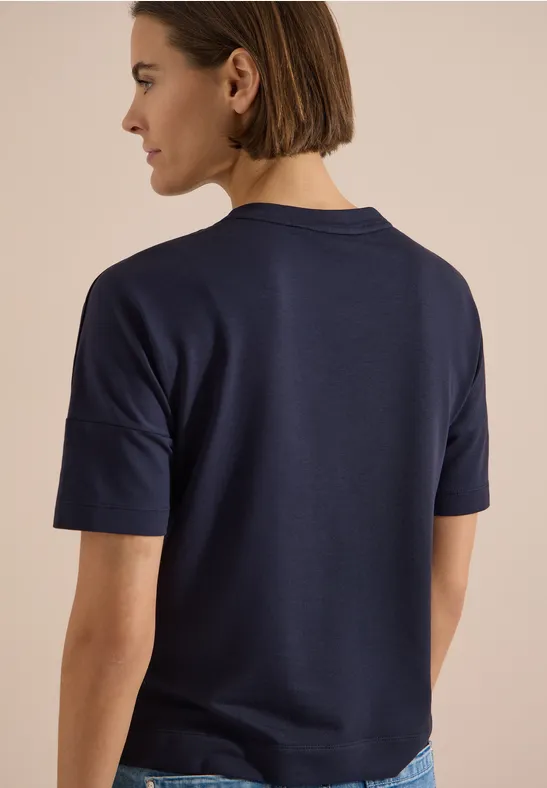Achteraanzicht van een vrouw in een donkerblauw T-shirt en spijkerbroek tegen een lichte achtergrond.
