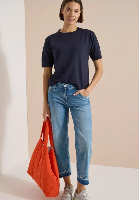 Persoon in donkerblauw T-shirt, lichtblauwe jeans en een oranje gewatteerde tas voor een beige achtergrond.