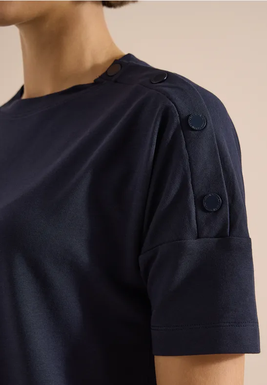 Vrouw in donkerblauw t-shirt met korte mouwen en decoratieve knopen op de schouder.