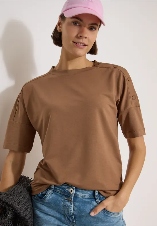 Personne en t-shirt marron à manches courtes avec boutons à l’épaule, jean bleu, une main dans la poche.