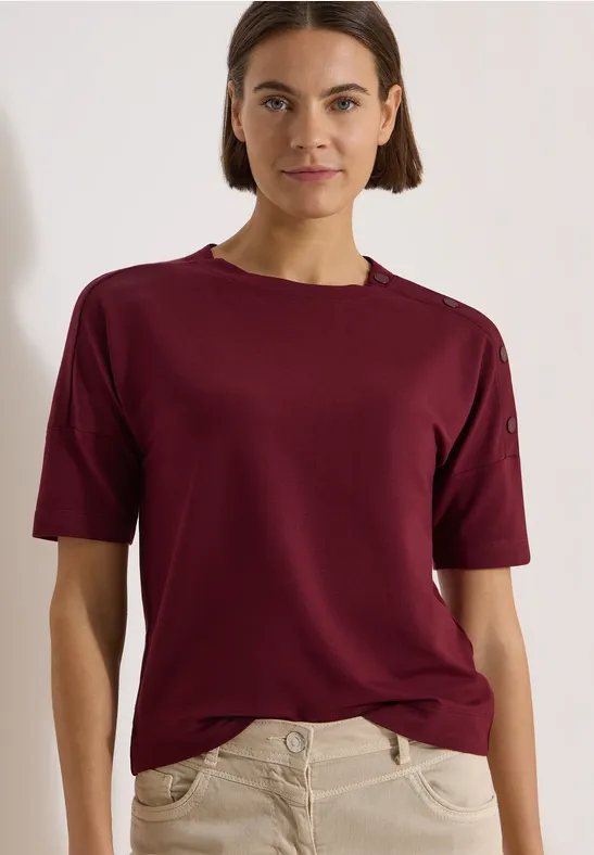 Une femme porte un t-shirt bordeaux à manches courtes et un pantalon clair devant un fond neutre.