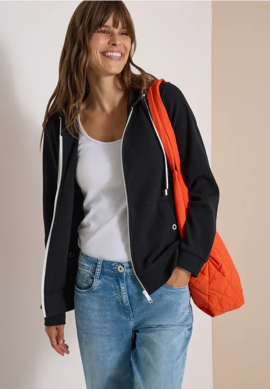 Een glimlachende vrouw in wit topje, zwart vest met capuchon, spijkerbroek en oranje tas.