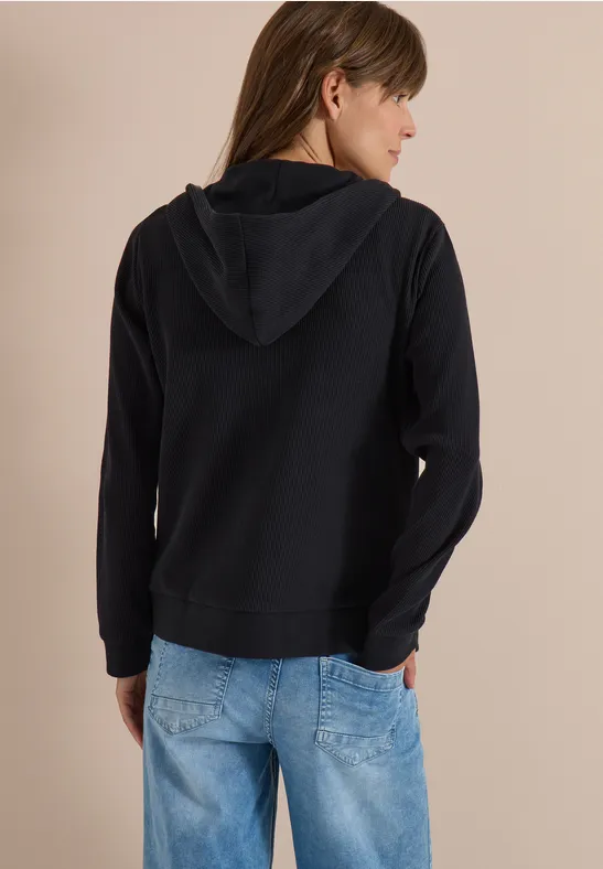Vrouw van achter gefotografeerd in een zwarte capuchontrui en lichtblauwe jeans voor een beige achtergrond.