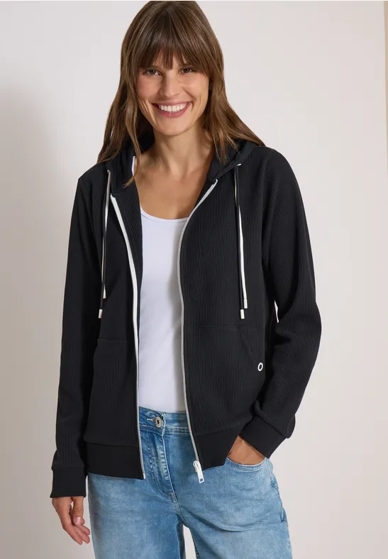 Een lachende vrouw draagt een zwarte capuchontrui over een wit shirt en blauwe jeans, neutrale achtergrond.