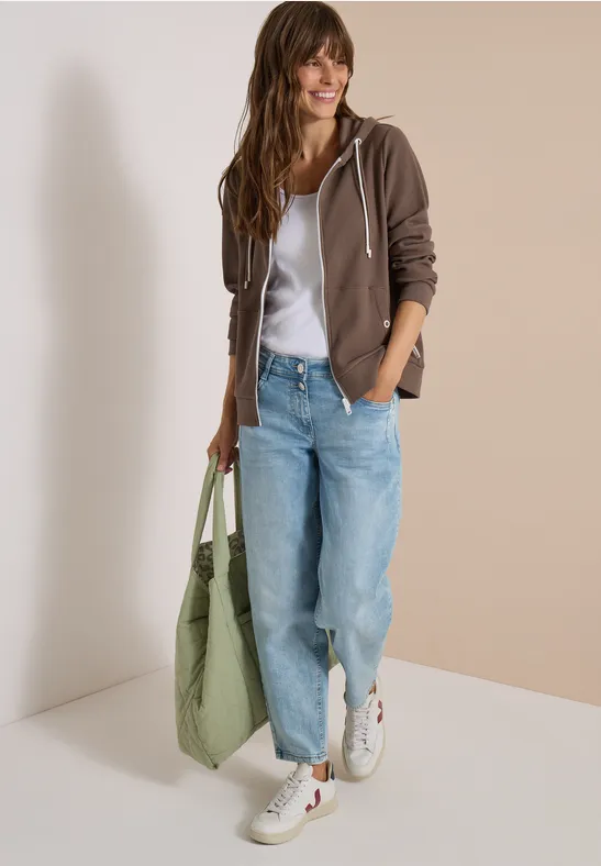 Vrouw in een casual look met een bruine capuchontrui, wit shirt, lichtblauwe jeans, witte sneakers en een groene tas.